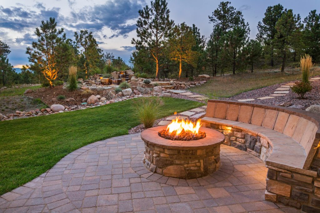 AdobeStock Image of a fire pit with a blazing fire surrounded by a nice bench, masonry patio, and grassy area.